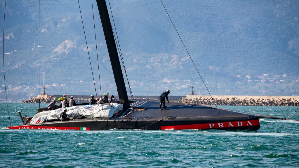 El AC75 "Luna Rossa Prada Pirelli" regresando al puerto, sobre un mar plano contra montañas en el fondo